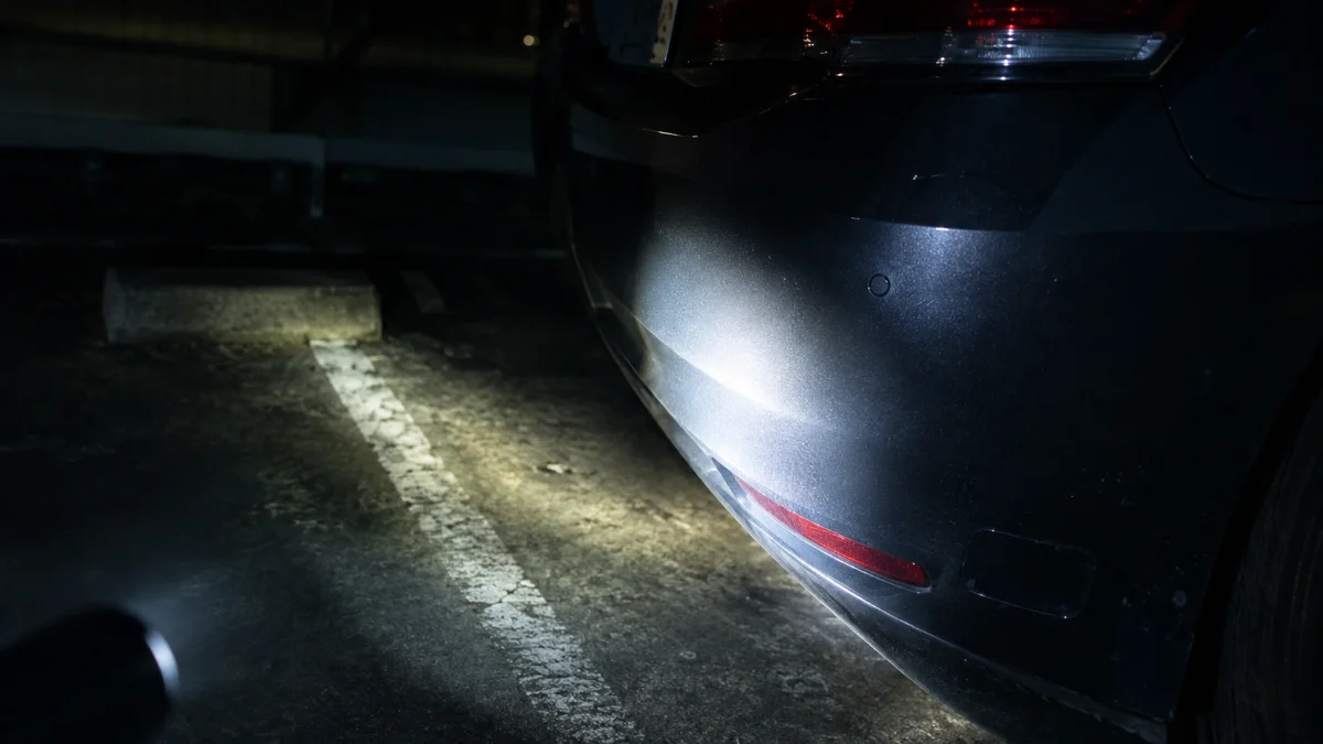 夜の駐車場に停められた車のバンパー付近を懐中電灯の光が照らしている様子