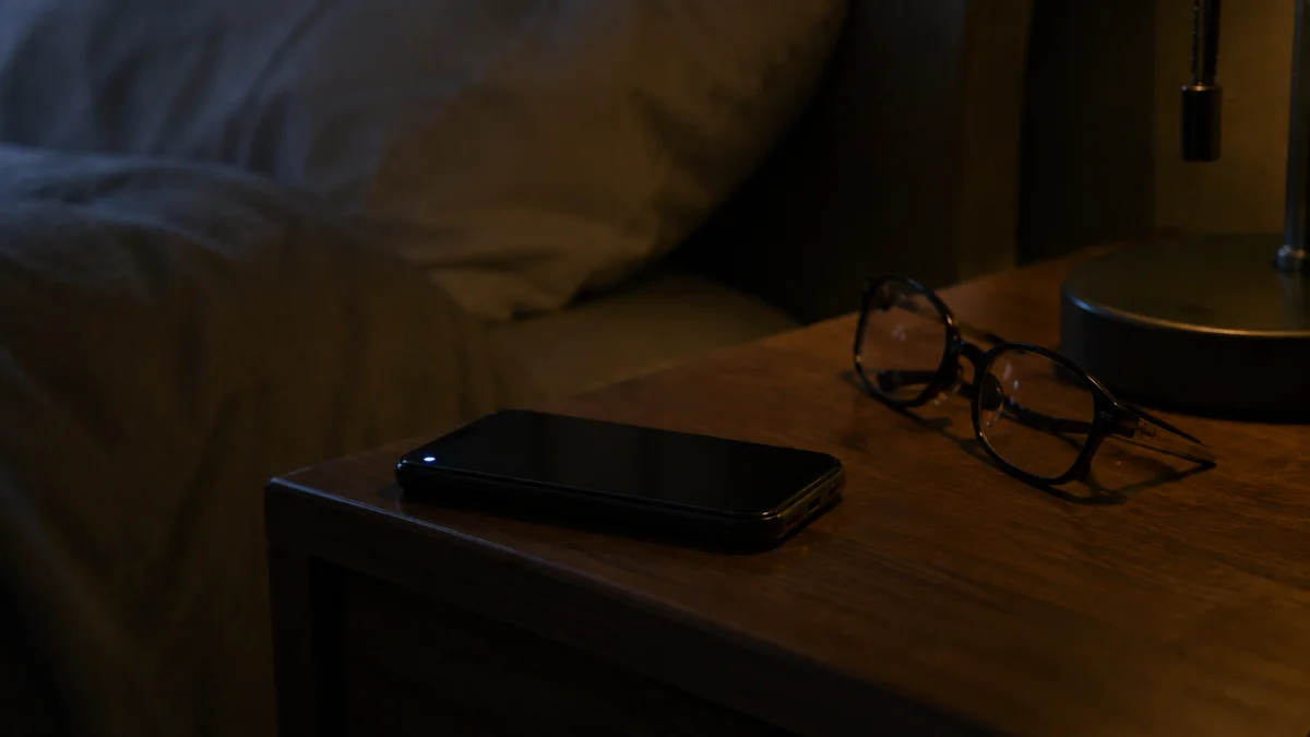 ナイトテーブルに置かれたスマートフォンと読書用の眼鏡、通知ライトが小さく光っている