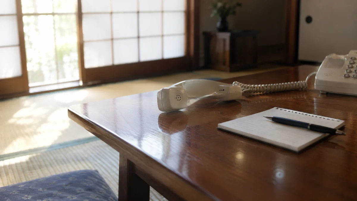 昼間の日本の和室で畳の上の座卓に置かれた固定電話の受話器と、その横のメモ帳とペン